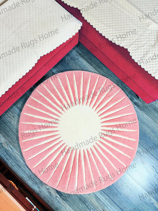 A round, hand-tufted rug with a sunburst design in pink and cream colors, placed on a wooden floor near red upholstered furniture with white quilted covers.
