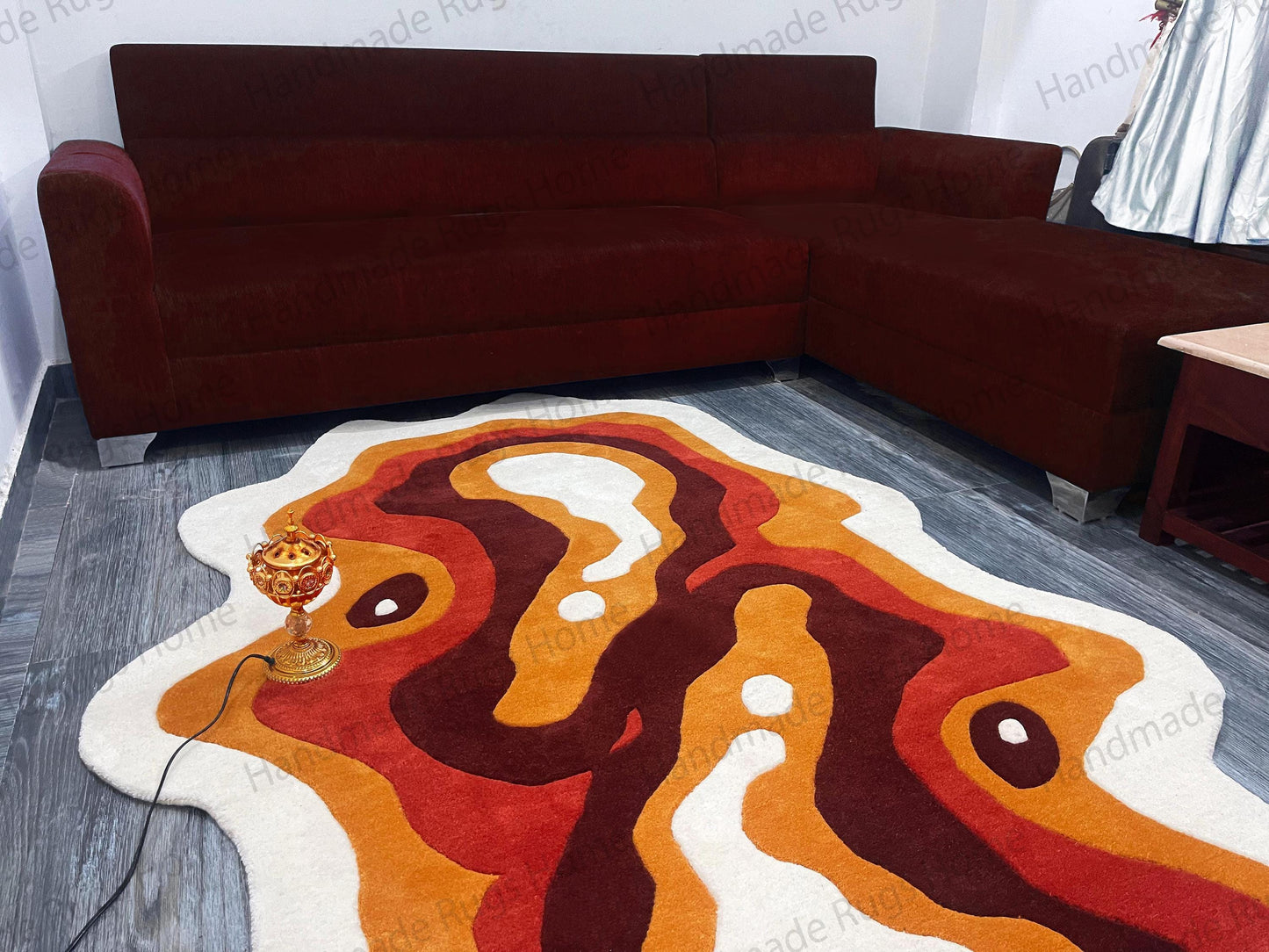A modern area rug with an abstract design. The rug features a flowing pattern of white, orange, red, and brown colors. A dark brown sectional sofa sits in the background. A gold decorative lamp sits on the rug.