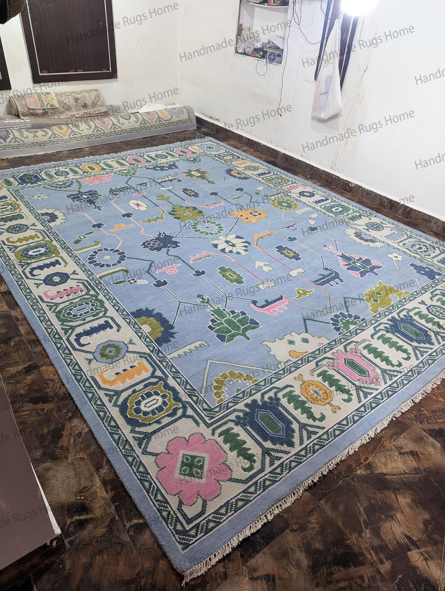 Hand-knotted (as per your request) rectangular rug with a light sky-blue background and a stylized, oversized floral design. Colors include blush pink, olive green, mustard, and navy blue. The rug features a heavy, ornate border and is displayed on a dark floor in a stockroom.