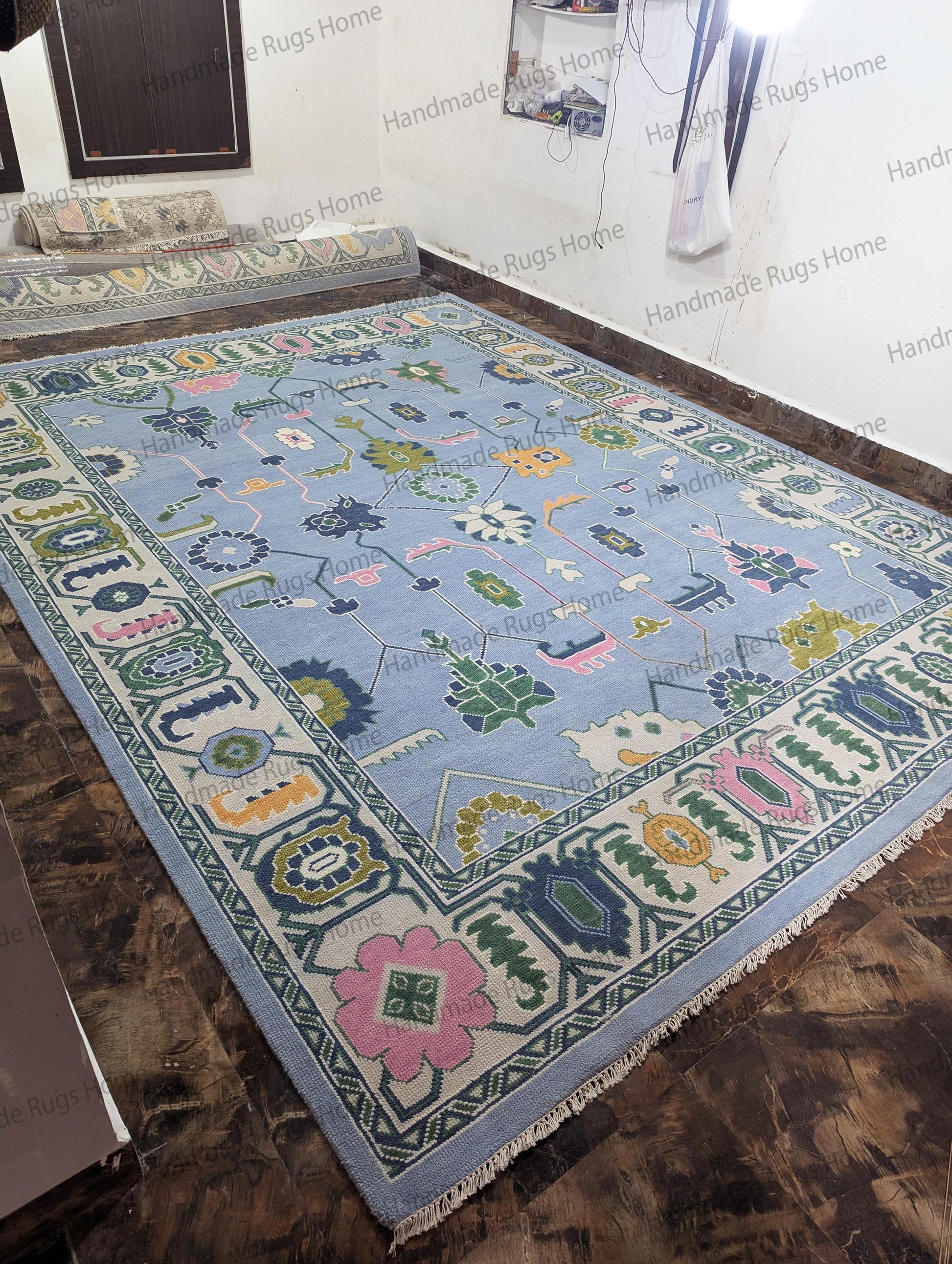 Hand-knotted (as per your request) rectangular rug with a light sky-blue background and a stylized, oversized floral design. Colors include blush pink, olive green, mustard, and navy blue. The rug features a heavy, ornate border and is displayed on a dark floor in a stockroom.